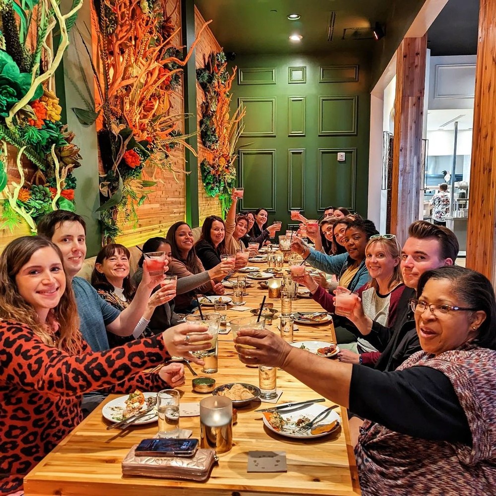 group of 18 people eating Dallas food at a restaurant. They are all raising a cocktail in celebration