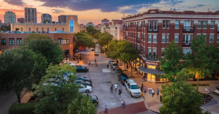 Street view of West Village in Dallas at sunset, featuring shops, restaurants, and pedestrians on a tree-lined avenue with historic buildings and modern architecture