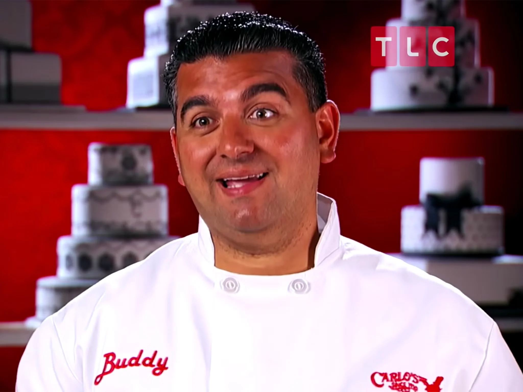 Celebrity chef Buddy Valastro, known for his TV show "Cake Boss" on TLC, wearing a white chef's jacket with his name embroidered on it, with decorated cakes in the background