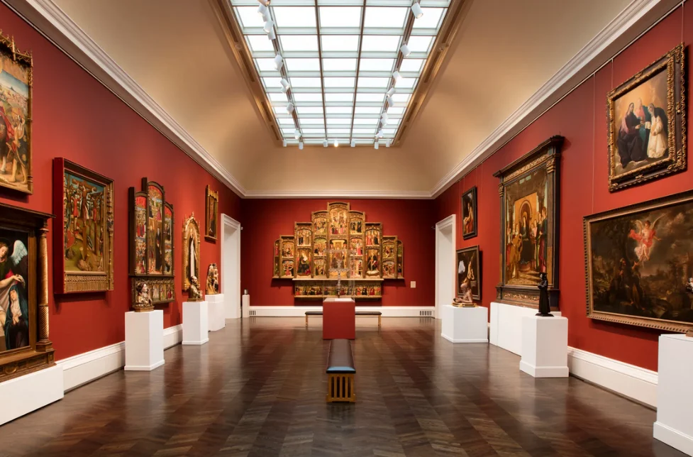 Art gallery in a Dallas museum featuring classical paintings and sculptures displayed on red walls with a skylight ceiling.