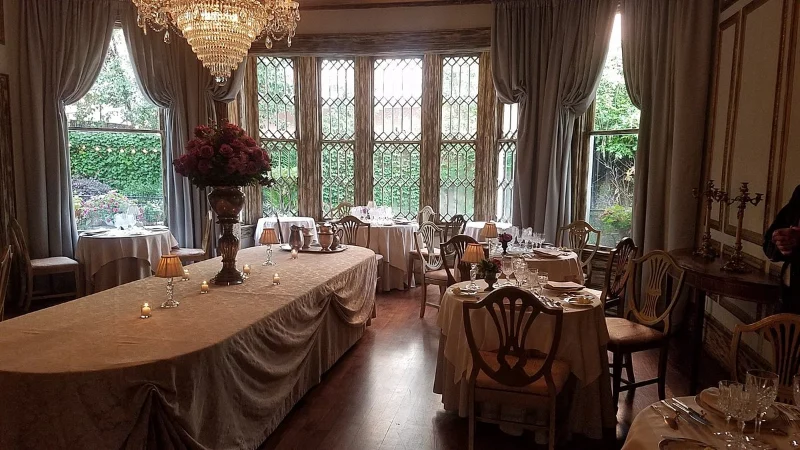 Elegant dining room with a large table set for a formal event, decorated with a chandelier, floral centerpiece, and surrounded by windows with garden views