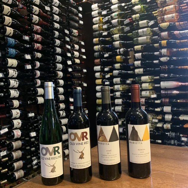 Four bottles of wine (OVR Old Vine Riesling, OVR Old Vine Red, Marietta ARMÉ Cabernet Sauvignon, Marietta ROMÁN Estate Grown) on a wooden table with a wall of wine bottles in the background