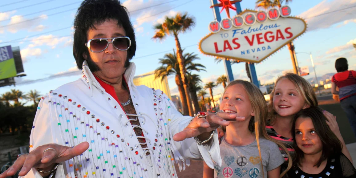 A man dressed as Elvis Presley poses with three young girls in front of the 'Welcome to Fabulous Las Vegas, Nevada' sign, with palm trees and a blue sky in the background.