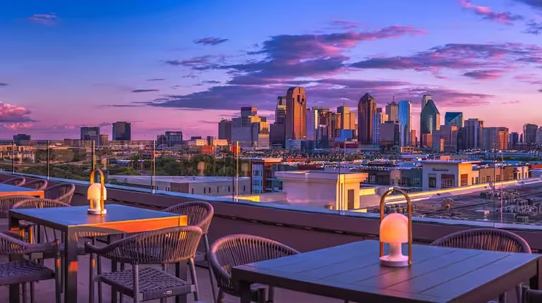 View of the Dallas skyline from the rooftop lounge of the Canopy Hotel at sunset, with outdoor seating and vibrant city lights