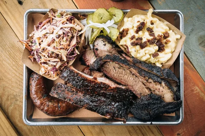 Plate of barbecue from a Dallas restaurant, featuring smoked sausage, brisket, ribs, coleslaw, pickles, and mac and cheese