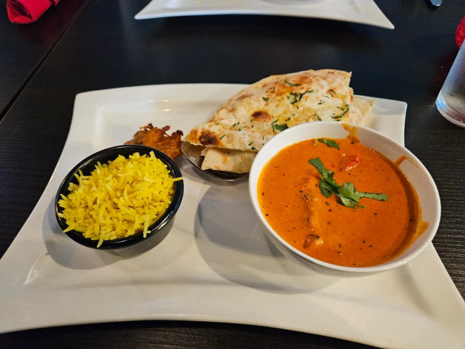 A plate of Indian cuisine featuring a bowl of creamy orange curry garnished with cilantro, a side of yellow rice, naan bread, and a small fritter, served on a white rectangular plate