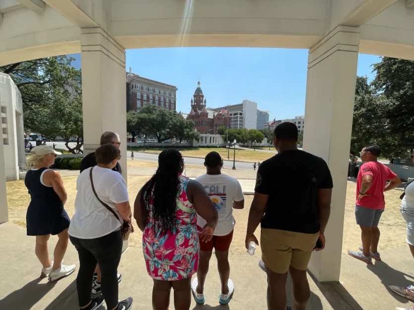 Group of people on a tour in downtown Dallas, standing under a pavilion and looking towards historic buildings and landmarks in the distance on a sunny day.