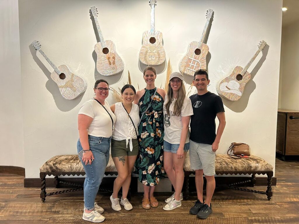 A group of people standing together inside a building at the Stockyards in Fort Worth, posing in front of a wall decorated with art pieces in the shape of guitars.